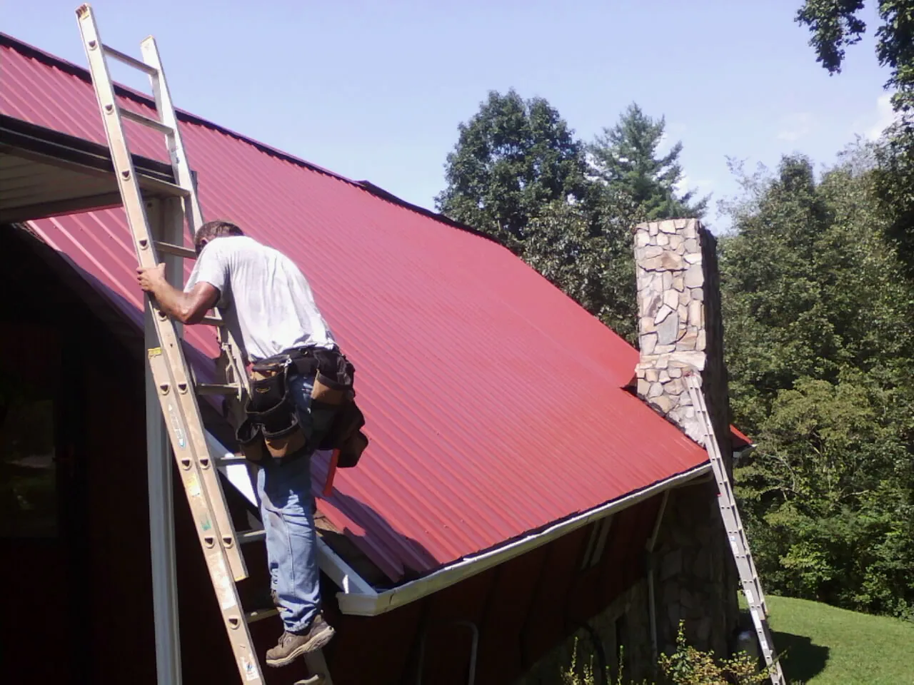 Roofer on a ladder beside a red metal roof and stone chimney — Soffit & Fascia Repair by Roofing Brothers in Port Jefferson Station